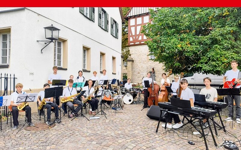 Die Schulbigband posiert auf dem Hemminger Marktplatz. Rechts im Bild die Rhythmusgruppe (Keyboards, Kontrabässe, E-Gitarre und Schlagzeug) auf der linken Seite die Bläser. Alle Schüler sind schick schwarz/weiß gekleidet. Im Hintergrund die Fachwerkarchitektur des Hemminger Stadtkerns.