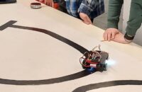 Ein selbstgebauter Roboter der Roboter-AG fährt auf einer aufgemalten Teststrecke, während Schüler:innen das Ergebnis beobachten.