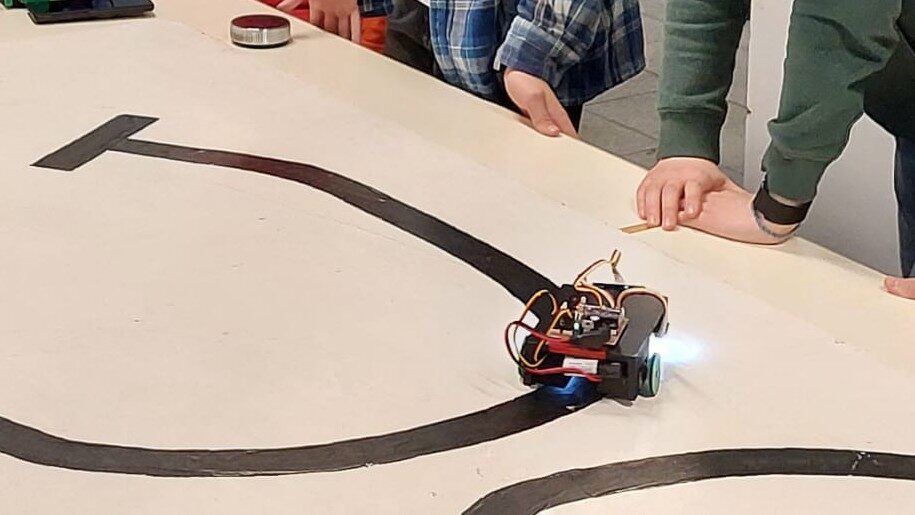 Titelbild-Roboter AG Ein selbstgebauter Roboter der Roboter-AG fährt auf einer aufgemalten Teststrecke, während Schüler:innen das Ergebnis beobachten.