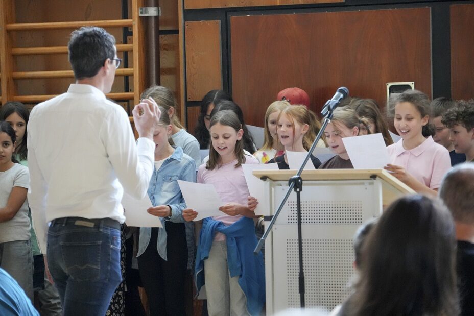 Der Chor der Fünftklässler singt ein selbstgedichtetes Begrüßungslied. Im Vordergrund von hinten gesehen Herr Strobel, ein Musiklehrer, dirigiert den Chor.