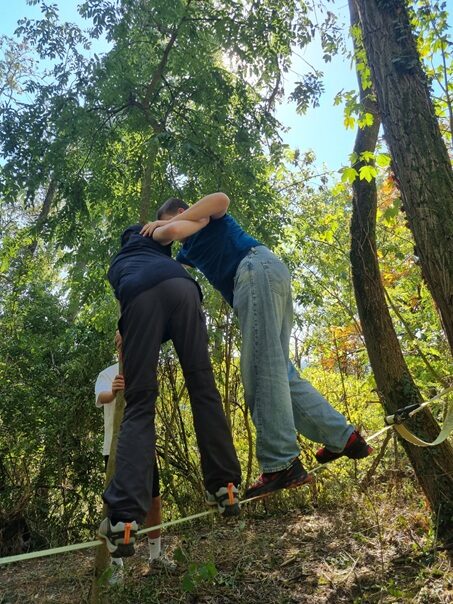Schüler der Klassenstuufe 9 helfen sich gegenseitig, eine Seilbrücke im Wald zu überqueren. Dei Schüler halten sich aneinander fest.