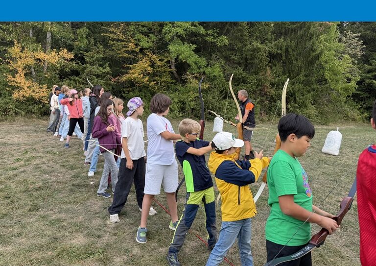 Kinder der 6c stehen in einer Reihe auf einer Wiese und üben, mit Pfeil- und Bogen zu schießen.