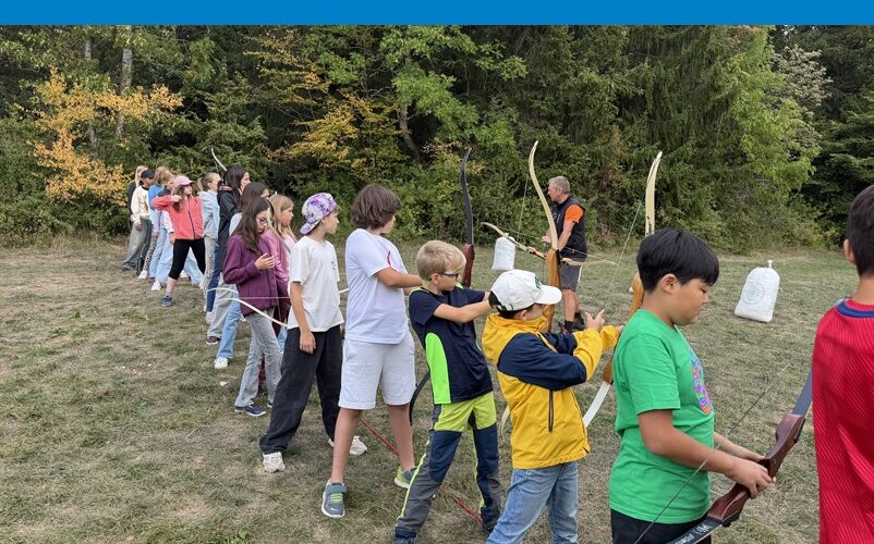 Kinder der 6c stehen in einer Reihe auf einer Wiese und üben, mit Pfeil- und Bogen zu schießen.